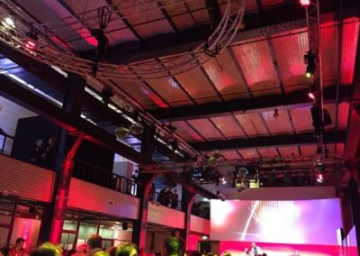 Blick auf Bühne, Publikum davor, Traversen mit Scheinwerfern, rotes Licht, große Leinwand über Bühne