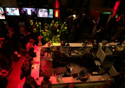 Blick von oben auf Bar, Leinwände mit Gesichtern und Bildern im Hintergrund, Menschen an Bar, Rote Beleuchtung