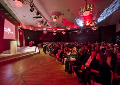 Publikum vor Bühne, rote Beleuchtung, Goldene Lampen und Lichtornamente an Decke, Bühne mit Rednerpult in Beige