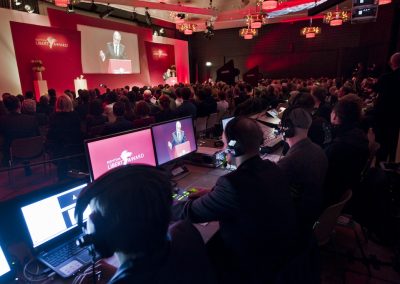 Blick aus Regie auf Bildschirme und Bühne auf der gerade ein Redner spricht