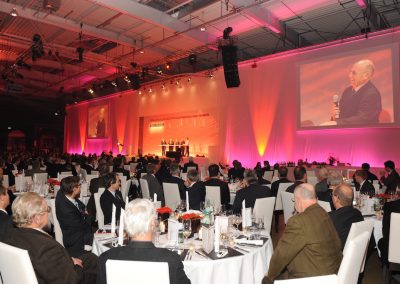 Gäste sitzen an runden Gala Tischen mit weissen Tischdecken und hören Redner auf Bühne zu, Bombardier, rotes Licht