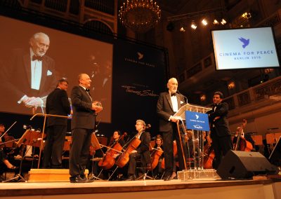 Mann in schwarzem Anzug mit Fliege spricht auf Bühne an Mikrofon, Orchester mit Instrumenten dahinter