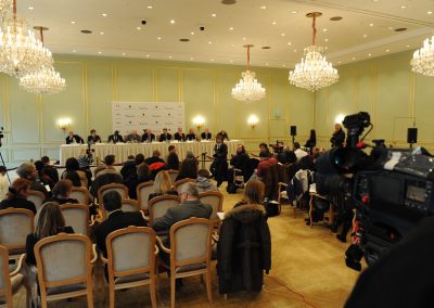 Pressekonferenz, weisse Kronleuchter und Sprecher auf der Bühne, Journalisten im Publikum