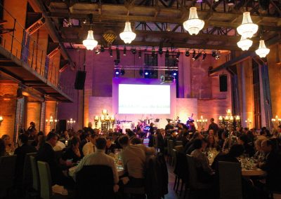 Gäste an Dinnertischen, große Kronleuchter an Decke, im Hintergrund Leinwand über Bühne