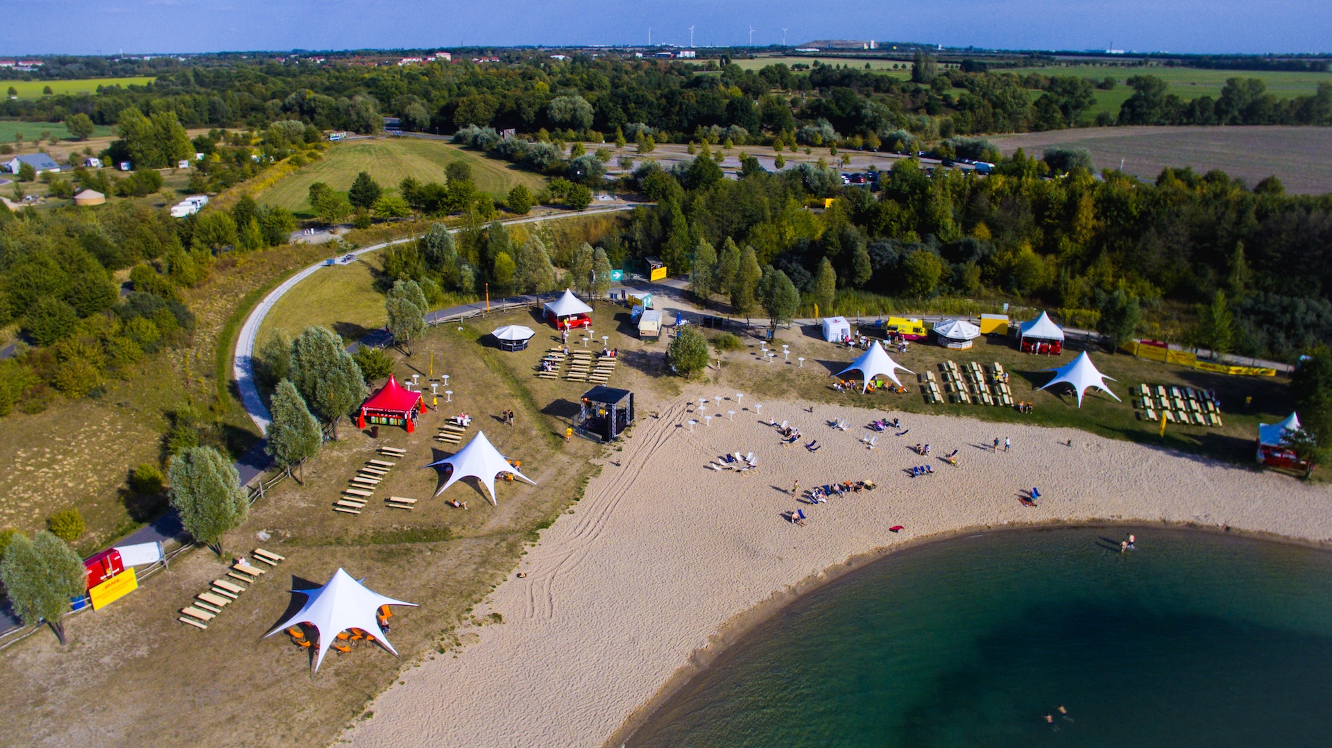 Schladitzer See mit Strand und Veranstaltungsfläche von oben, mit Zelten Bühne und Sitzgelegenheiten.