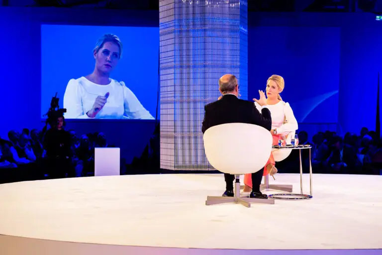 Hybrid Veranstaltung Zwei Personen in Interviewsituation auf runder Bühne sitzend umgeben von Publikum, abgefilmt und im Hintergrund groß projeziert