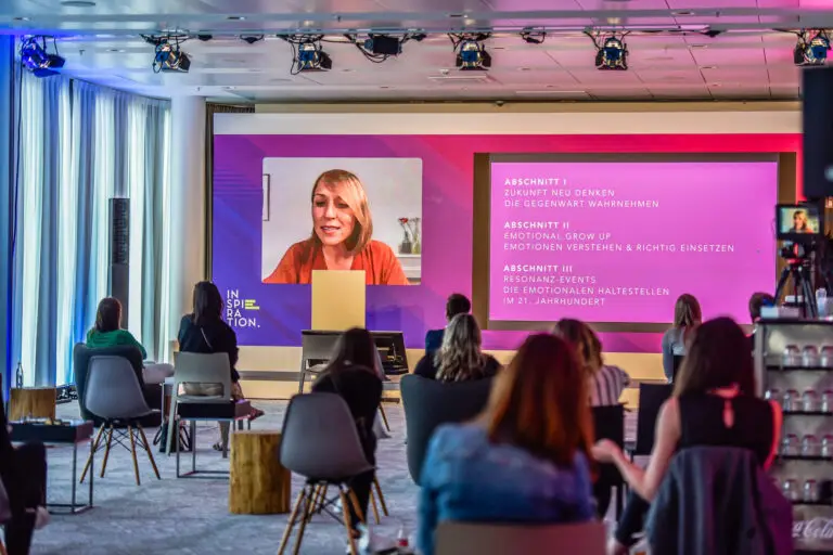 Hybride Veranstaltung im Konferenzformat, Vortragende am Raumende auf Großleinwand mit Konferenzagenda zugeschaltet, Anwesende Teilnehmer auf Stühlen davor
